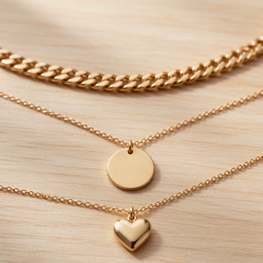 A chunky gold curb chain and two fine gold-tone pendant chains with a disc and a heart charm, laid out in parallel on a warm wood surface