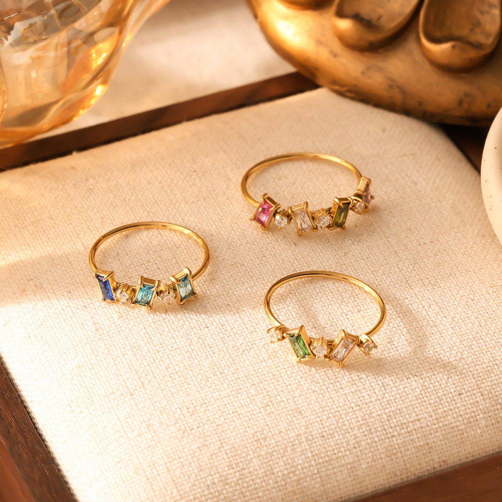 Four small gold-tone stainless steel rings with coloured cubic zirconia stones on a reflective gold surface