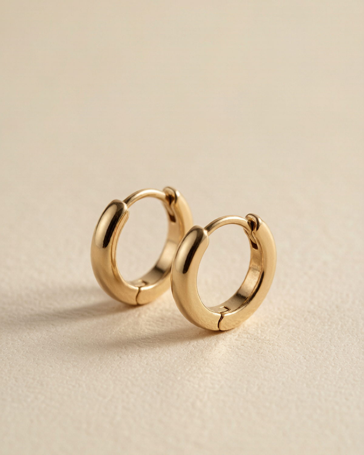 A pair of small gold-tone huggie earrings with visible hinge closures, photographed on a warm beige background
